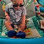 baby, sunglasses, play_mat, toy, indoor, child, blue_shoes, shirt, star_pattern, floor, colorful, curious, seated, hand, head, face, clothing, footwear, baby_toys, playtime