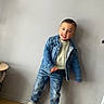 child, toddler, denim_jacket, jeans, sneakers, smile, standing, indoor, wooden_floor, wall, table, playful, happy, casual_clothing, portrait, person, fashion, cute, young, light