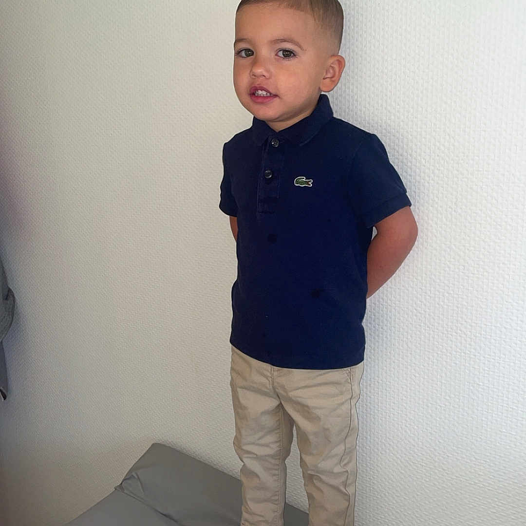 Noam participe au concours pour gagner de l'argent avec cette photo : child, boy, standing, navy_shirt, polo_shirt, beige_pants, white_sneakers, indoor, wall, padded_surface, casual_clothing, short_hair, footwear, looking, person, young, portrait, standing_pose, minimal_background, confident