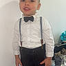 Noam a rejoint le concours — aidez-le/la à gagner de superbes lots ! child, boy, formal_clothing, bow_tie, suspenders, white_shirt, pants, standing, indoor, smiling, portrait, person, face, hair, hand, furniture, bottle, household_items, casual, background