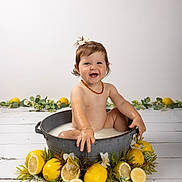 Jade participe au concours pour gagner de l'argent avec cette photo : baby, child, smiling, milk_bath, metal_tub, lemons, fruit, flowers, greenery, white_background, wooden_floor, cute, playful, hair_bow, jewelry, bracelet, portrait, sitting, happy, indoors