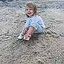 beach, casual_clothing, child, daylight, footprints, grass, happy, nature, outdoor, person, playful, portrait, rocks, sand, shoe, sitting, small_rocks, smiling, toddler, young_child
