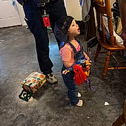 Sage is registered to the contest to win money with this photo: adult, bag, cap, casual_wear, chair, child, clothing, colorful, concrete_floor, doll, expression, floor, gift_bag, home, indoor, looking_up, overalls, person, standing, toy