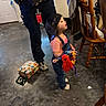 adult, bag, cap, casual_wear, chair, child, clothing, colorful, concrete_floor, doll, expression, floor, gift_bag, home, indoor, looking_up, overalls, person, standing, toy