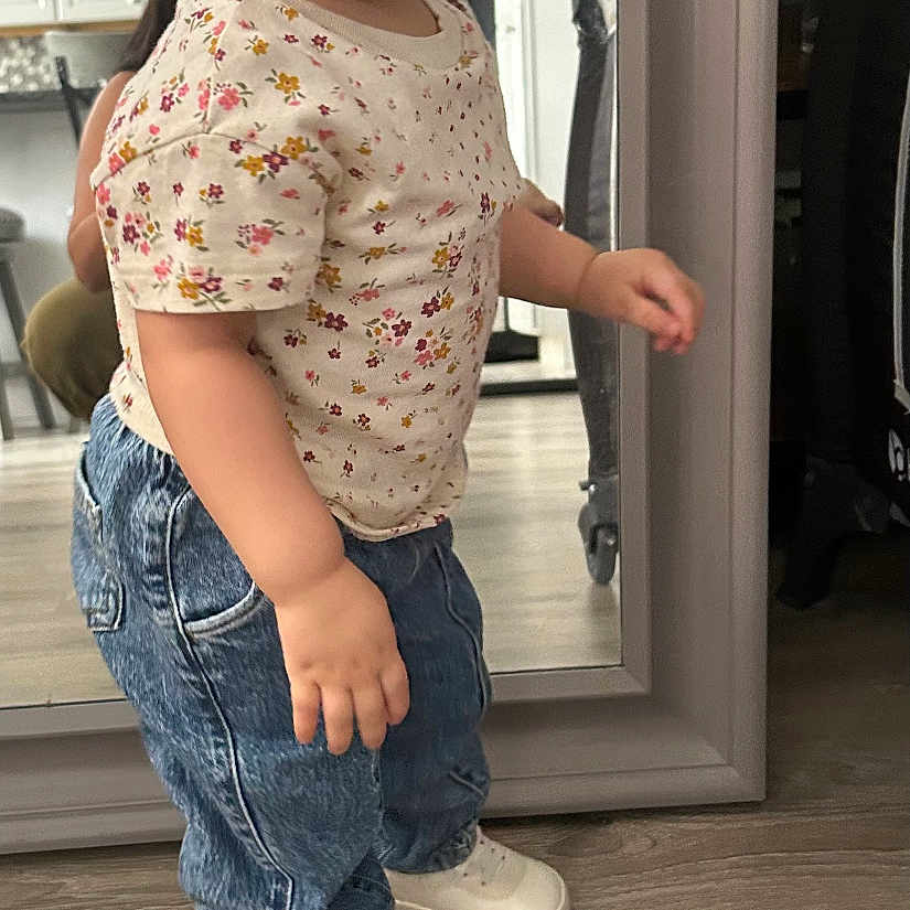 Arleth is registered to the contest to win money with this photo: toddler, child, mirror, headband, jeans, sneakers, indoor, flooring, reflection, person, white_shirt, wood_floor, cabinet, curious, standing, casual_clothing, hair_accessory, home, portrait, real_photo