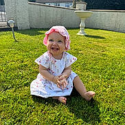 Mila is registered to the contest to win money with this photo: baby, child, smiling, grass, outdoor, sunlight, pink_hat, white_dress, happy, clapping, garden, nature, summer, cute, sitting, footwearless, greenery, daytime, wall, residential