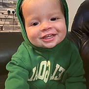 Hudson is registered to the contest to win money with this photo: child, toddler, hoodie, green_clothing, smiling, face, indoor, living_room, sofa, clock, wall, framed_pictures, happy, cute, person, sweatshirt, young_child, candid, portrait, cozy