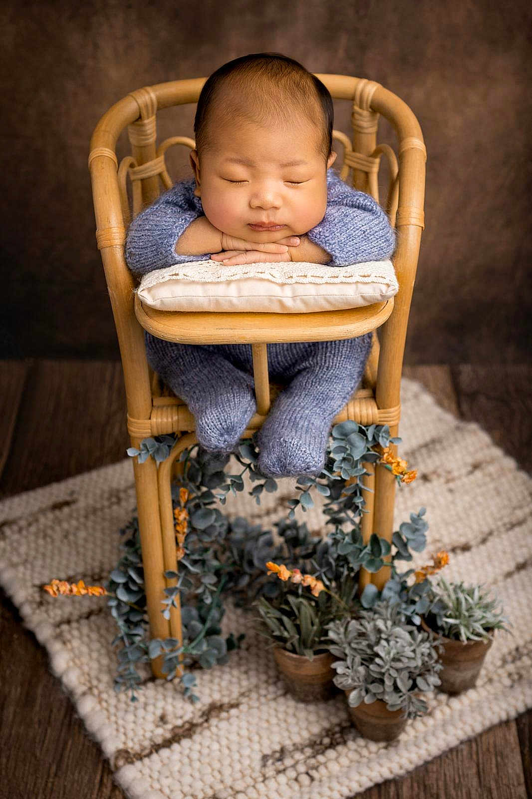 Sangsehang is registered to the contest to win money with this photo: baby, sleeping, blue_knit_outfit, wooden_chair, pillow, potted_plants, greenery, indoor, cozy, peaceful, child, portrait, floor_rug, decor, relaxation, infant, knitwear, hands, face, head