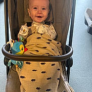 Caden is registered to the contest to win money with this photo: baby, infant, child, stroller, smiling, happy, portrait, face, sitting, blanket, toy, bird_toy, indoor, sunlight, harness, carriage, fabric, brown, colorful, family