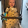 baby, infant, child, stroller, smiling, happy, portrait, face, sitting, blanket, toy, bird_toy, indoor, sunlight, harness, carriage, fabric, brown, colorful, family