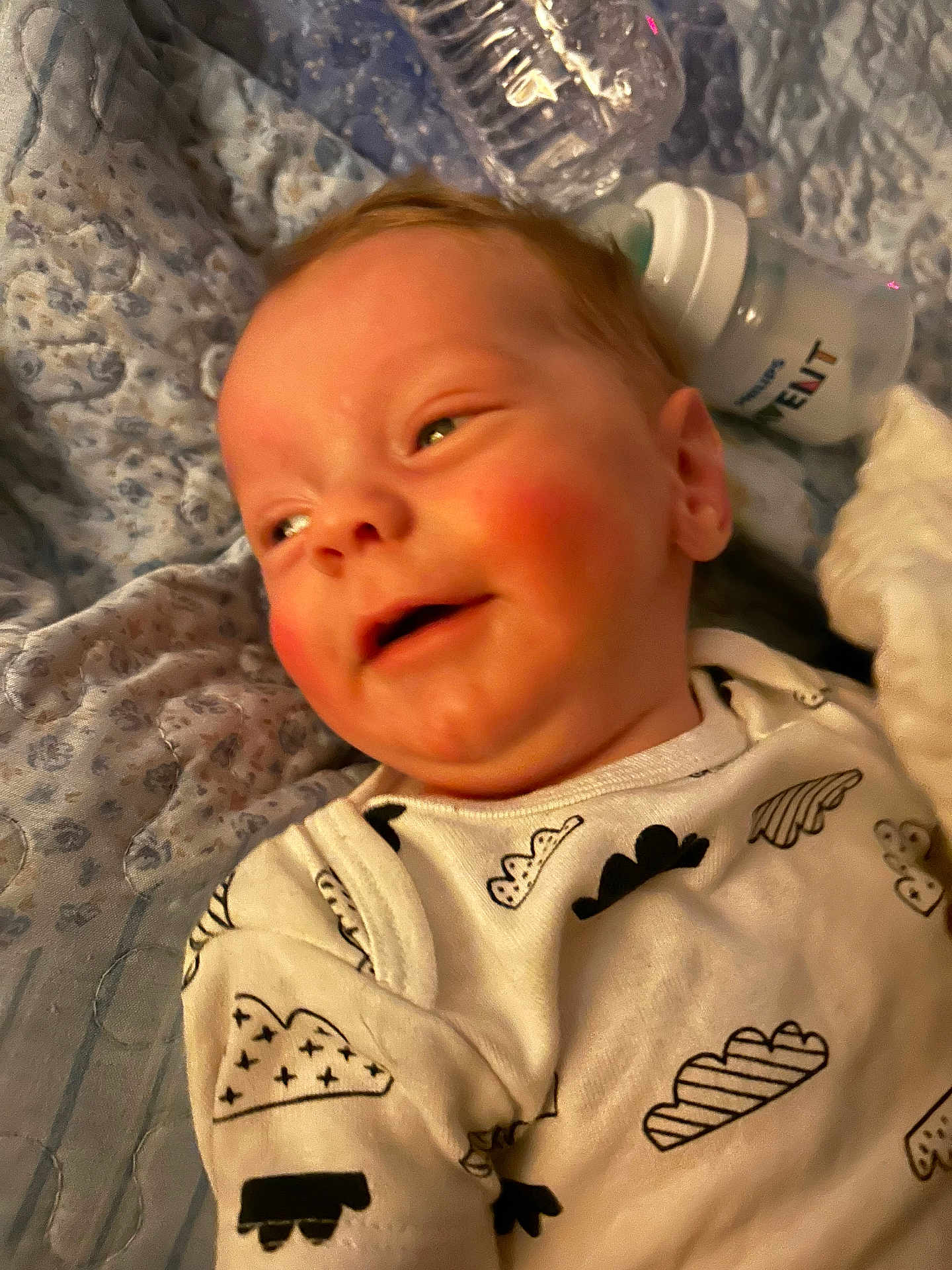 Tristan joined the competition — help win amazing prizes! baby, infant, smiling, onesie, cloud_pattern, blanket, quilt, bottle, baby_bottle, water_bottle, face, cheeks, lying_down, closeup, cute, child, newborn, soft_lighting, indoor, portrait