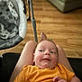 Tristan is registered to the contest to win money with this photo: baby, child, infant, orange_shirt, person, lap, home_interior, wooden_floor, baby_swing, furniture, relaxing, casual, indoor, cute, smiling, skin, head, hands, legs, seated