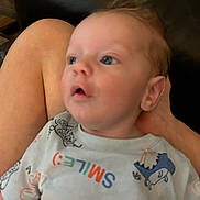 Tristan is registered to the contest to win money with this photo: baby, infant, child, person, clothing, shirt, dinosaur, smile_text, sitting, lap, skin, head, face, eyes, mouth, hand, leg, background, indoor, blurred