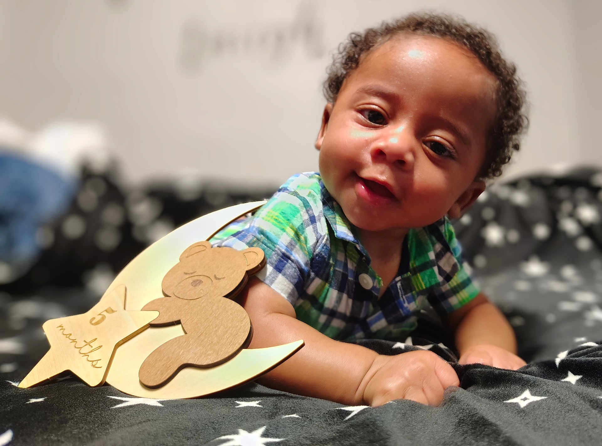 Octavious is registered to the contest to win money with this photo: baby, infant, child, plaid_shirt, smiling, lying_down, blanket, star_pattern, wooden_decoration, bear, 5_months, cute, indoors, portrait, face, hand, curly_hair, cozy, soft_lighting, closeup