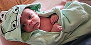 Vincent is registered to the contest to win money with this photo: baby, infant, towel, green_towel, frog_hood, hooded_towel, wrapped, hands, face, eyes, indoor, carpet, wooden_cabinet, legs, skin, comfort, cozy, person, resting, cute