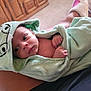 baby, infant, towel, green_towel, frog_hood, hooded_towel, wrapped, hands, face, eyes, indoor, carpet, wooden_cabinet, legs, skin, comfort, cozy, person, resting, cute