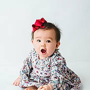 Parker joined the competition — help win amazing prizes! baby, infant, toddler, bow, hair_bow, floral_dress, patterned_clothing, sitting, barefoot, toes, open_mouth, surprised_expression, portrait, studio, white_background, cute, adorable, long_sleeves, one_person, hands
