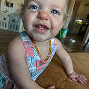 Camilla is registered to the contest to win money with this photo: toddler, child, smile, blue_eyes, floral_dress, amber_necklace, close_up, hands, cushion, indoor, home, flooring, furniture, person, cute, happy, portrait, baby, skin, blond_hair