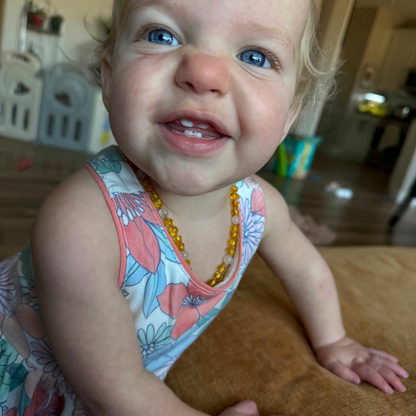Camilla is registered to the contest to win money with this photo: toddler, child, smile, blue_eyes, floral_dress, amber_necklace, close_up, hands, cushion, indoor, home, flooring, furniture, person, cute, happy, portrait, baby, skin, blond_hair