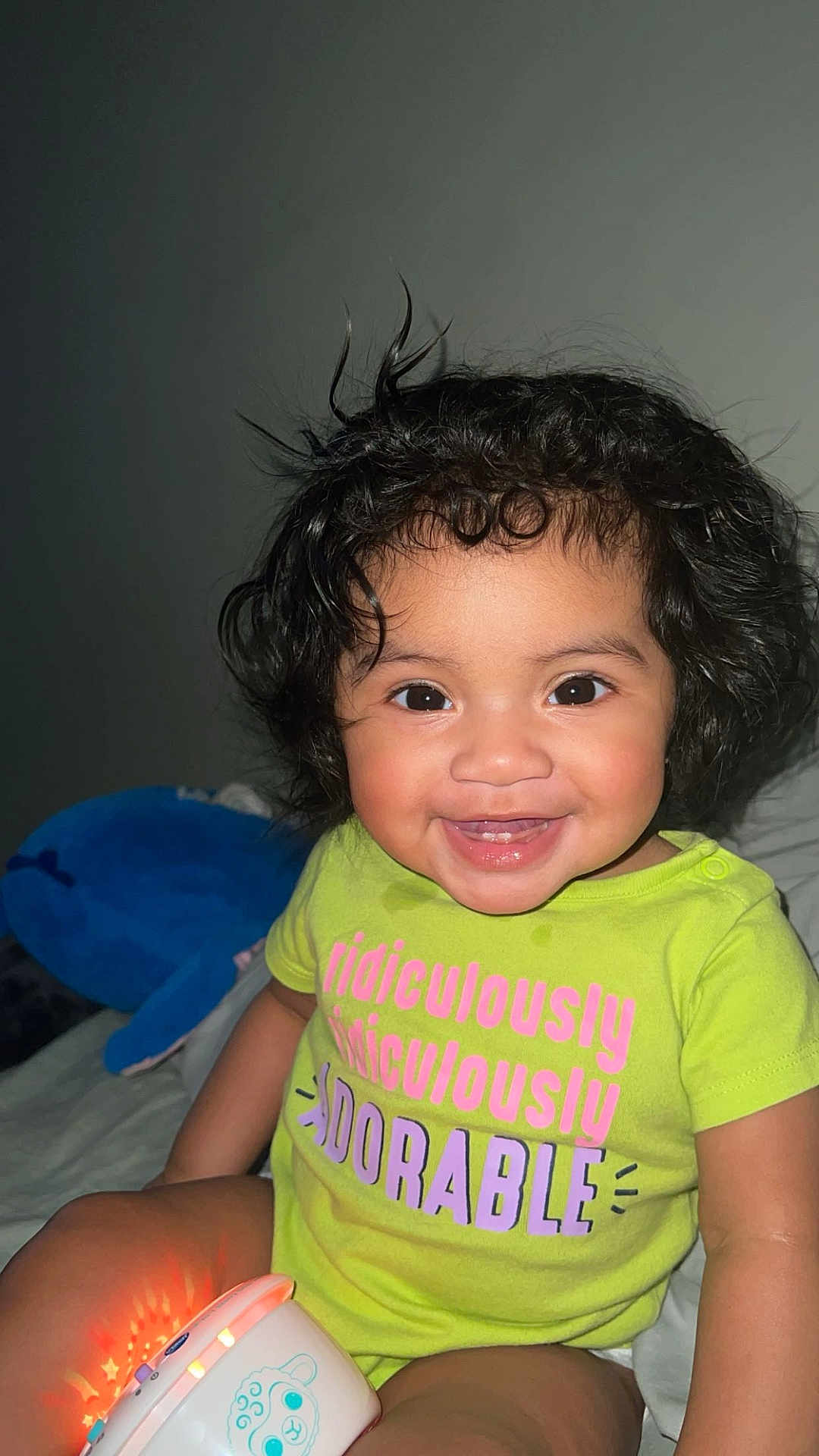 Maddison is registered to the contest to win money with this photo: toddler, child, smiling, curly_hair, bright_eyes, green_shirt, text_on_shirt, toy, glowing_toy, bed, plush_toy, blue_stuffed_animal, indoors, happy, cute, portrait, baby, person, sitting, fun