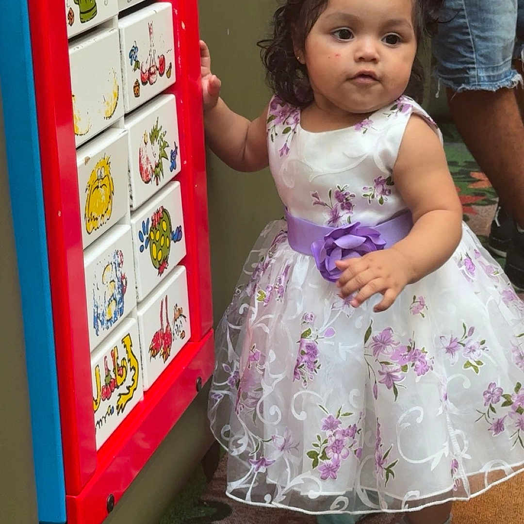 Ciara is registered to the contest to win money with this photo: toddler, child, girl, dress, purple_bow, playground, play_panel, carpet, curious, indoor, person, footwear, sock, shorts, adult, pattern, floral, colorful, toy, exploring