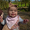 Odie is registered to the contest to win money with this photo: baby, child, smiling, headband, pink_bib, outdoor, forest, trees, sunlight, person, happy, cute, nature, daylight, holding, infant, portrait, clothing, hand, dirt_path