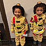 child, toddler, firefighter_costume, toy_axe, smiling, curious, indoor, wooden_floor, textured_wall, costume, black_shoes, curly_hair, yellow_outfit, red_stripes, play, role_play, two_children, standing, young, portrait