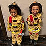 child, toddler, firefighter_costume, toy_axe, smile, laughing, curly_hair, matching_outfits, indoor, wall, floor, shoes, play, costume, hat, reflection_strips, emotion, standing, person, fun