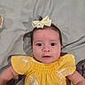 Luna is registered to the contest to win money with this photo: baby, infant, child, yellow_dress, bow, head_accessory, lying_down, bed_sheet, curious_expression, face, cute, indoors, portrait, young_child, soft_lighting, human, skin, arm, eye_contact, closeup