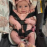 baby, child, car_seat, pink_clothing, headband, smiling, infant, safety_harness, cute, indoor, footwear, pacifier, floral_pattern, seatbelt, portrait, person, baby_girl, happy, hand, closeup