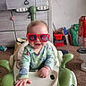 Penelope is registered to the contest to win money with this photo: baby, child, sunglasses, toy_car, indoor, smiling, green, clothing, furniture, car_seat, play, happy, cute, infant, carpet, cabinet, toy, red, person, home