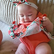 Penelope is registered to the contest to win money with this photo: baby, infant, child, orange_outfit, headband, floral_pattern, cloth, cushion, texture, skin, cute, indoors, sitting, curious, soft_light, portrait, person, hand, legs, closeup