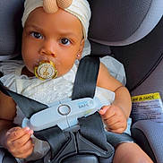 Leilany is registered to the contest to win money with this photo: baby, child, car_seat, pacifier, headband, flower, white_clothing, safety_harness, seatbelt, infant, cute, portrait, indoors, child_safety, serious_expression, toddler, cute_accessory, straps, car_interior, small_hands