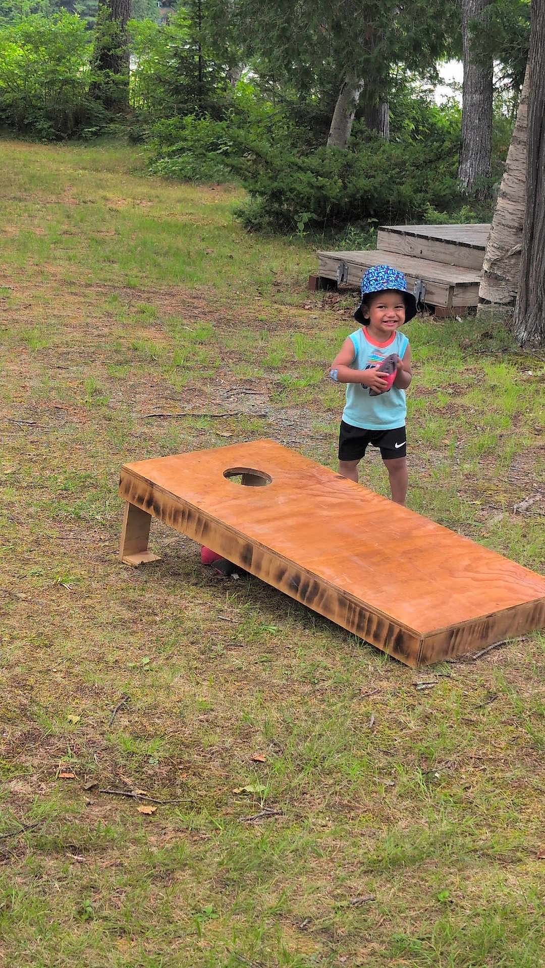 Zehri is registered to the contest to win money with this photo: child, outdoor, grass, sport, game, cornhole, wooden_board, football, hat, smile, happy, nature, trees, deck, summer, play, casual_clothing, young, fun, recreation
