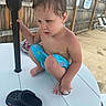 toddler, child, boy, swim_trunks, bare_chest, table, pole, crocs, footwear, outdoor, fence, concrete, poolside, chairs, summer, curious, crouching, hair, skin, daylight