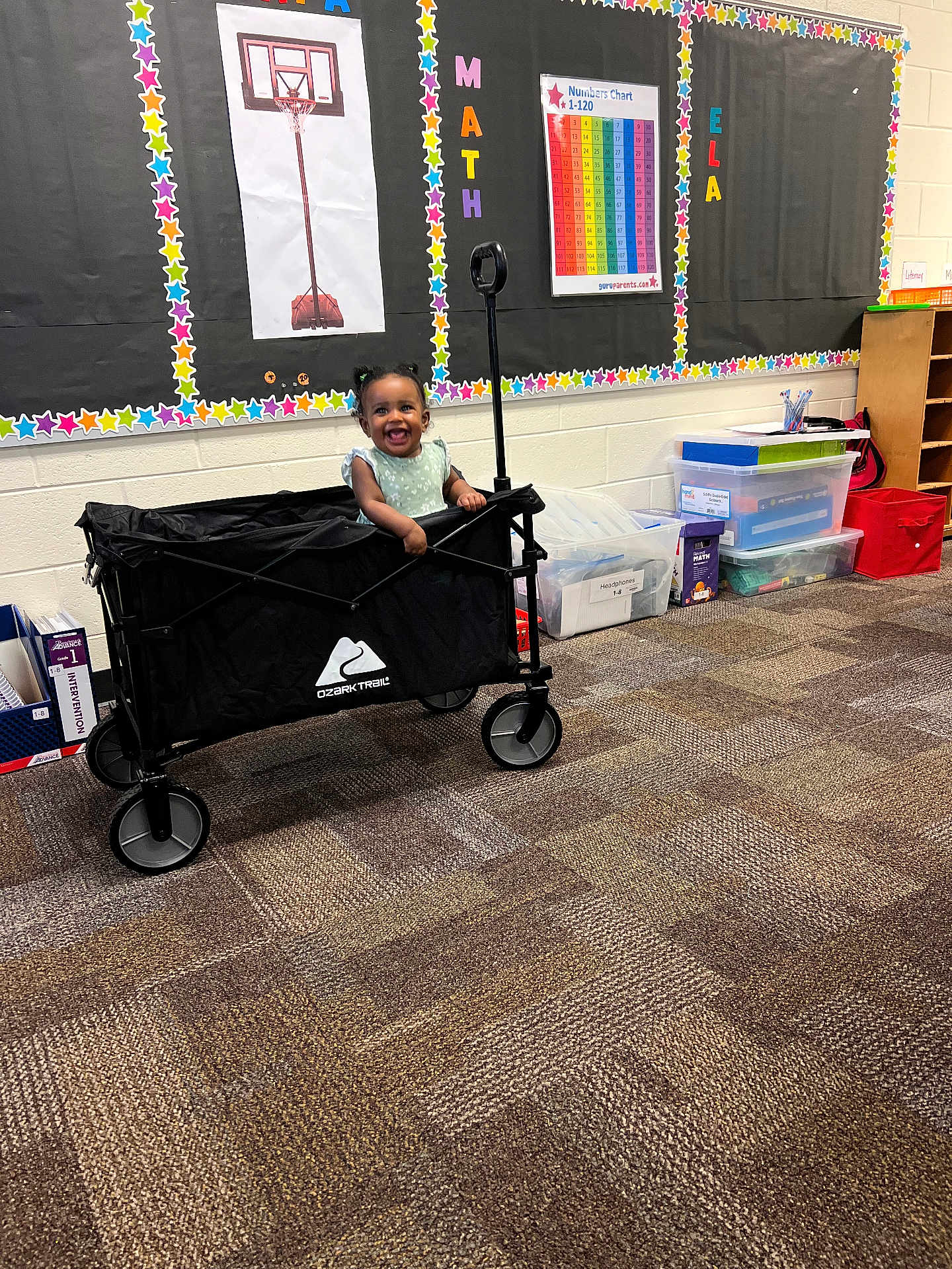 Alaya is registered to the contest to win money with this photo: toddler, child, wagon, classroom, carpet, posters, storage_bins, smile, happy, educational, indoor, toy, person, colorful, basketball_poster, numbers_chart, floor, handle, hair_buns, wall