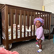 Alaya joined the competition — help win amazing prizes! toddler, child, crib, wooden_crib, purple_bonnet, polka_dot_clothing, smiling, standing, carpet, stuffed_toy, blanket, bedroom, natural_light, shadow_pattern, cozy, furniture, indoor, happy, person, baby