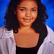 Dakota is registered to the contest to win money with this photo: child, portrait, smiling, curly_hair, dark_hair, face, eyes, headshot, studio, blue_background, white_shirt, black_top, clothing, person, looking_at_camera, front_facing, shoulders, indoor, photography, confident_expression