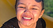 Dakota joined the competition — help win amazing prizes! child, smiling, laughing, portrait, close_up, face, eyes_closed, teeth, curly_hair, dark_hair, earrings, black_shirt, sparkly_clothing, yellow_chair, outdoor, greenery, sunlight, happy, joyful, photography