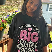 Dakota is registered to the contest to win money with this photo: child, girl, smiling, portrait, curly_hair, black_shirt, big_sister, outdoor, porch, flowers, potted_plants, garden, house, window, balcony, daylight, family, fashion, happy, standing