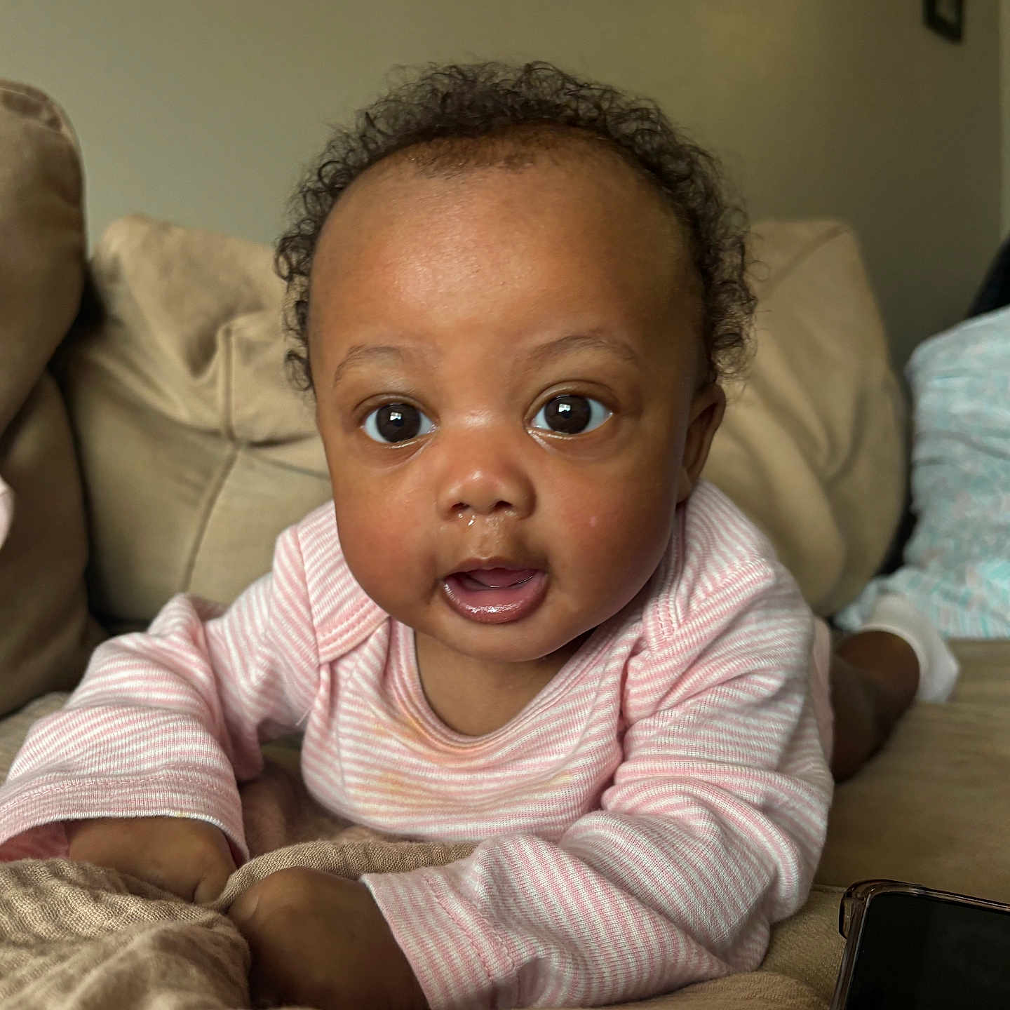 Sayge is registered to the contest to win money with this photo: baby, infant, cute, face, eyes, pink_clothing, striped_clothing, couch, living_room, curly_hair, skin, child, indoors, portrait, expression, person, toddler, blanket, home, cozy