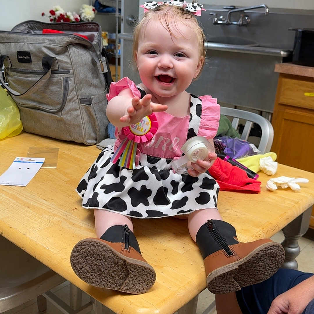 Cheyanne is registered to the contest to win money with this photo: baby, bag, boots, bow, cabinet, child, dress, floor, furniture, hand, happy, indoor, kitchen, person, playful, ribbon, sitting, smiling, table, tape