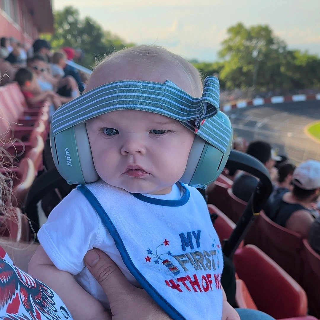Joshua joined the competition — help win amazing prizes! baby, child, headphones, bib, tattoo, adult_hand, stadium_seating, outdoor, racetrack, blue_sky, people, plaid_shorts, bandage, person, summer, event, spectator, daytime, cute, patriotic