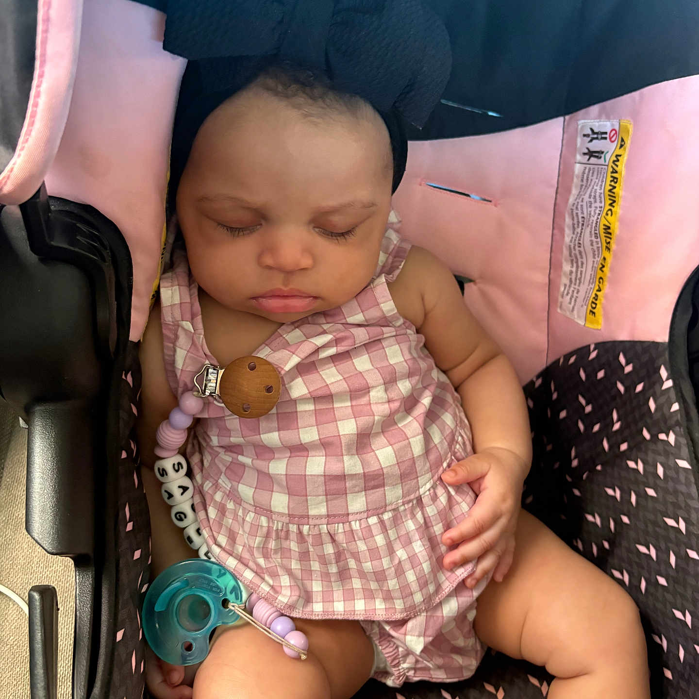 Sagelyn is registered to the contest to win money with this photo: baby, sleeping, car_seat, pacifier, headband, bow, pink, checkered, clothing, child, infant, cute, peaceful, resting, seat, strap, accessory, indoors, person, portrait