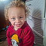 casual_clothing, child, clutter, curly_hair, drawer, face, floor, happy, indoor, light, mickey_mouse, person, plastic_furniture, portrait, red_hoodie, smiling, toddler, toy, window, young
