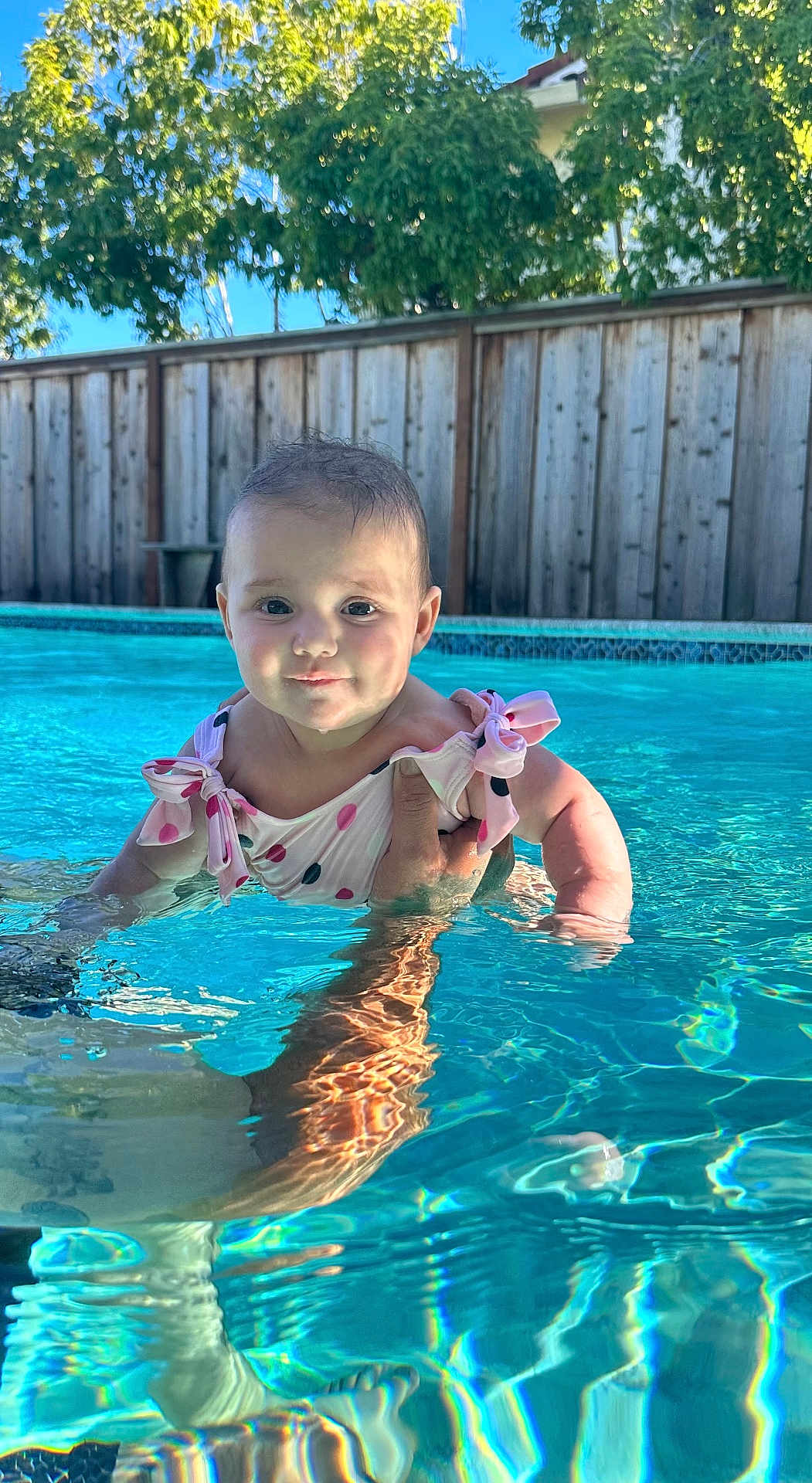 Anastasia is registered to the contest to win money with this photo: baby, child, swimming_pool, water, summer, outdoor, sunlight, reflection, polka_dot, swimsuit, cute, smiling, person, hand, skin, trees, fence, blue, daytime, recreation