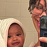 baby, towel, smiling, glasses, adult, child, mirror, bathroom, selfie, person, face, skin, eyes, hand, portrait, curly_hair, closeup, indoor, happy, holding