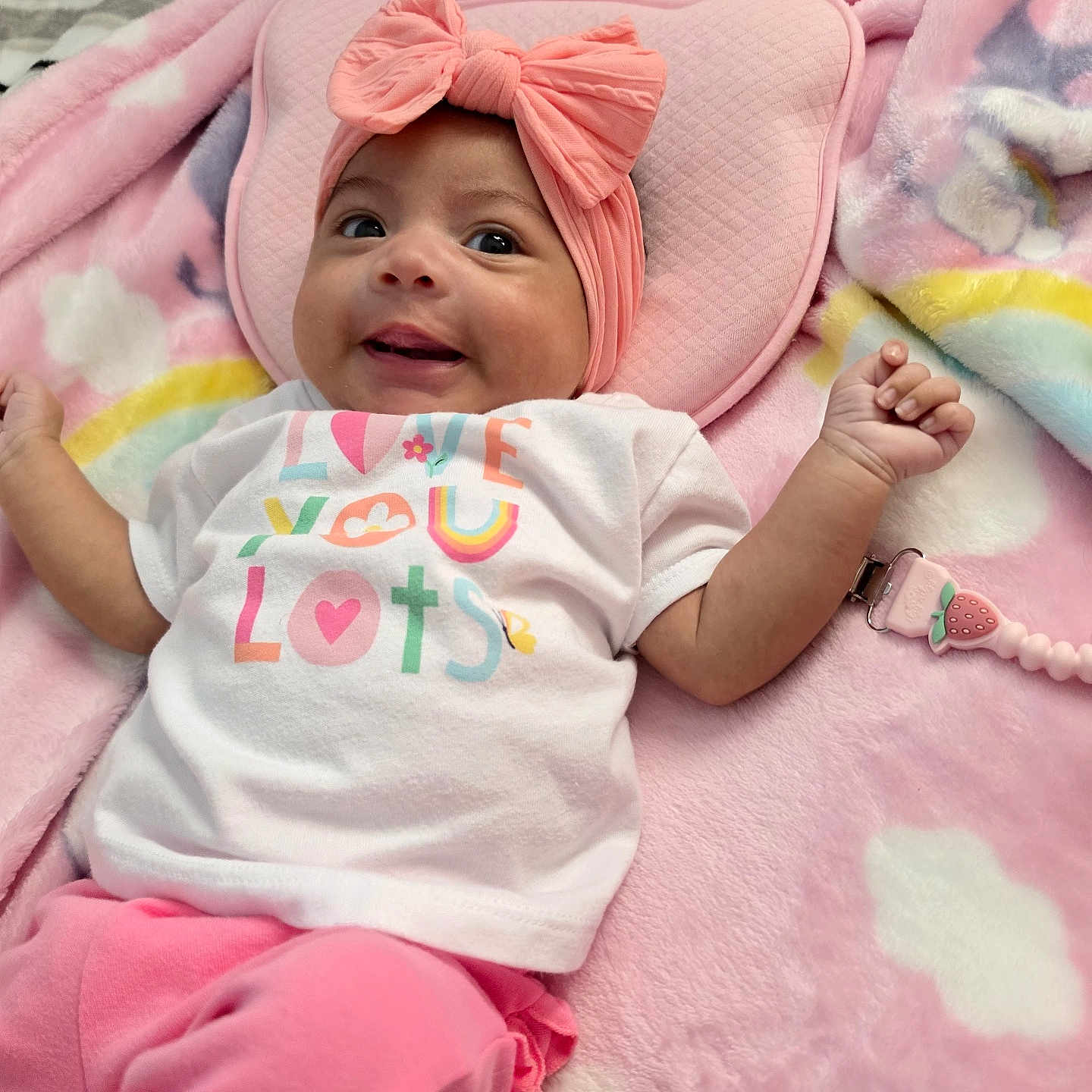 Sofia is registered to the contest to win money with this photo: baby, infant, child, smiling, portrait, pink, bow, headband, onesie, clothing, blanket, bed, lying_down, toy, pacifier, face, arms, indoor, colorful, happy