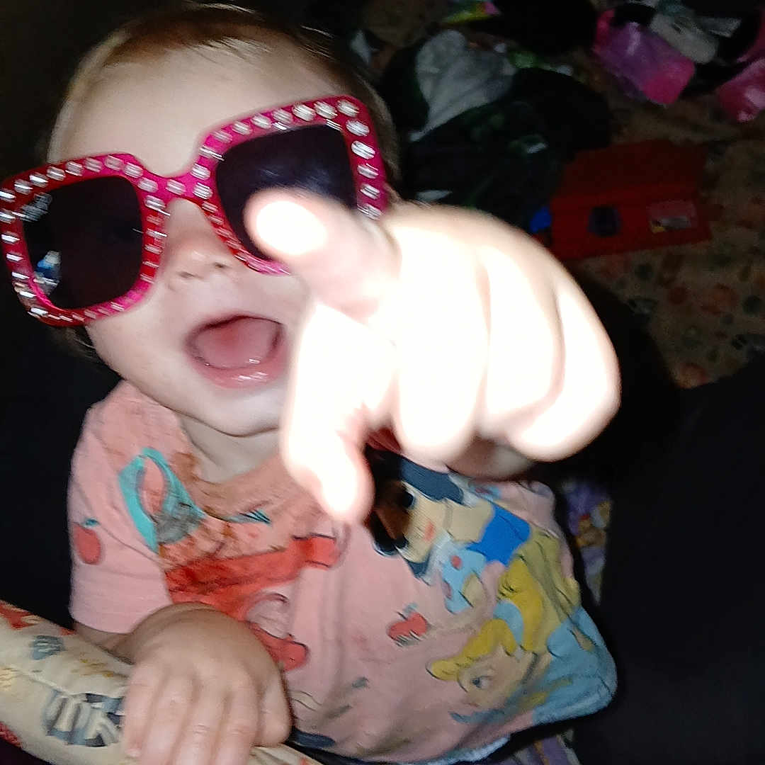 Kinlinn is registered to the contest to win money with this photo: background, casual, child, clothing, colorful, expression, face, fun, hand, indoors, joyful, padded_rail, person, pink, playful, pointing, smiling, sunglasses, toddler, toy