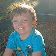 Dylan is registered to the contest to win money with this photo: child, smiling, boy, outdoor, sunlight, blue_tshirt, dinosaur_graphic, grass, leaves, happy, portrait, casual_clothing, nature, daylight, young_child, face, head, short_hair, playful, park